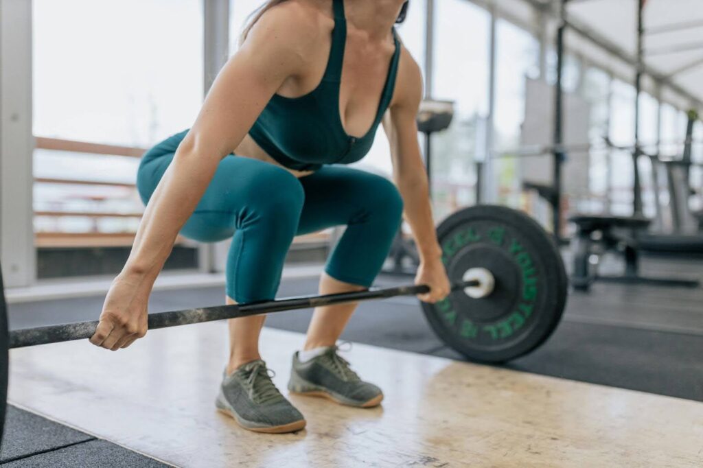 Close-up of a female athlete in teal workout clothes preparing to deadlift a barbell in the gym, symbolizing the strength and performance needs of endurance athletes and highlighting the risks of training fatigue and female overtraining symptoms when cutting out carbs without proper recovery nutrition.